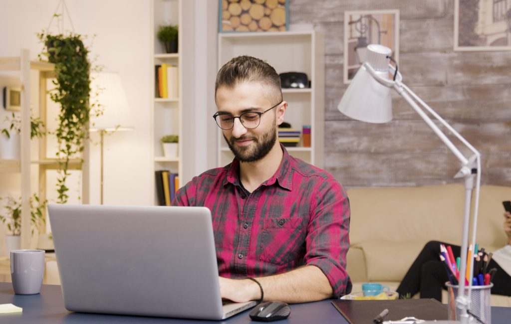 Handsome man laughing while working on laptop in living room. Girlfriend in the background talks on the phone.