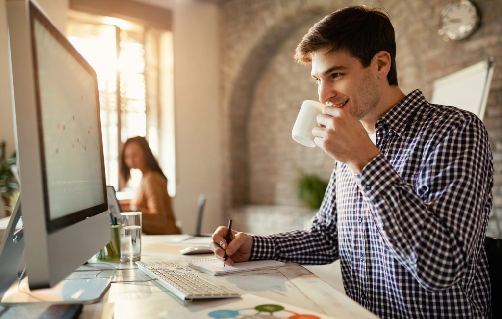 Smiling businessman drinking coffee while taking notes and using desktop PC in the office.